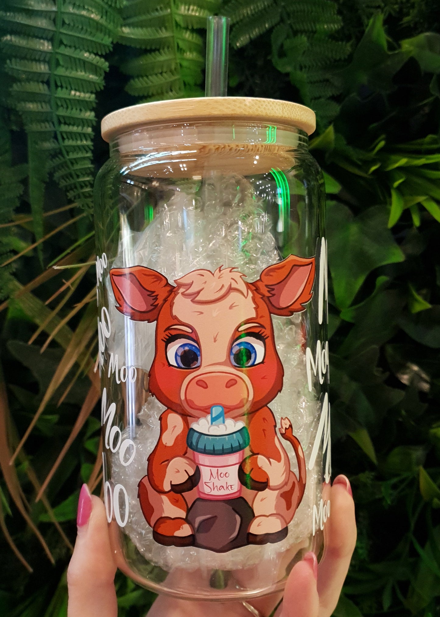 Clear tumbler with cartoon cow design held by a hand against a green leafy background