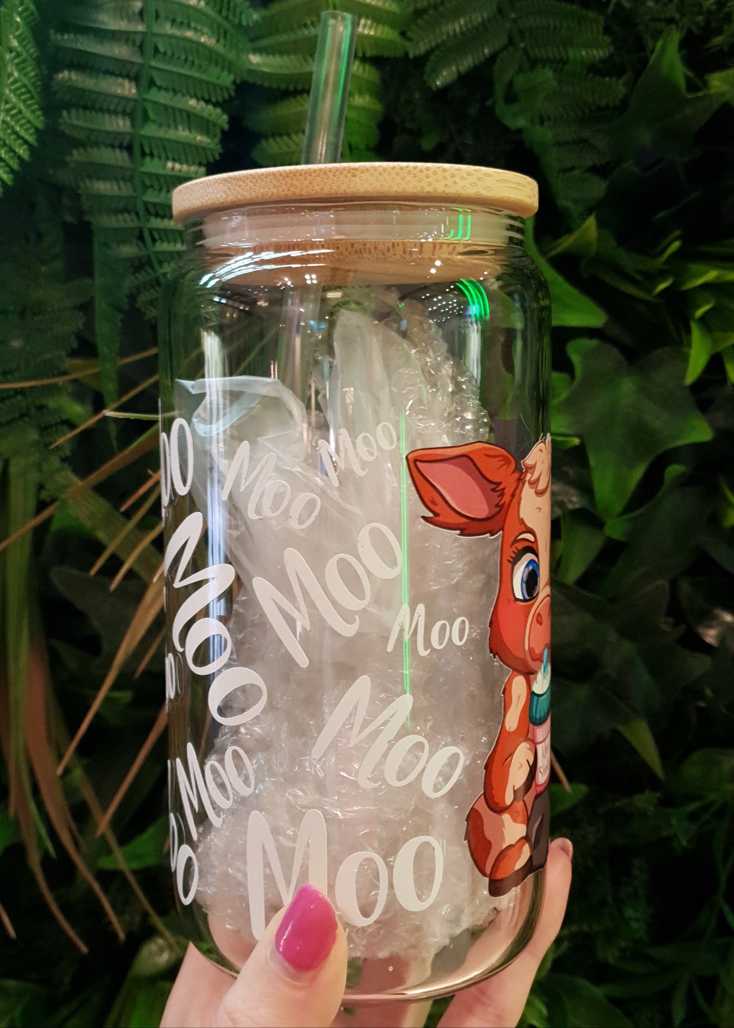 Clear tumbler with cow design and 'Moo' text held by a hand on green leaves
