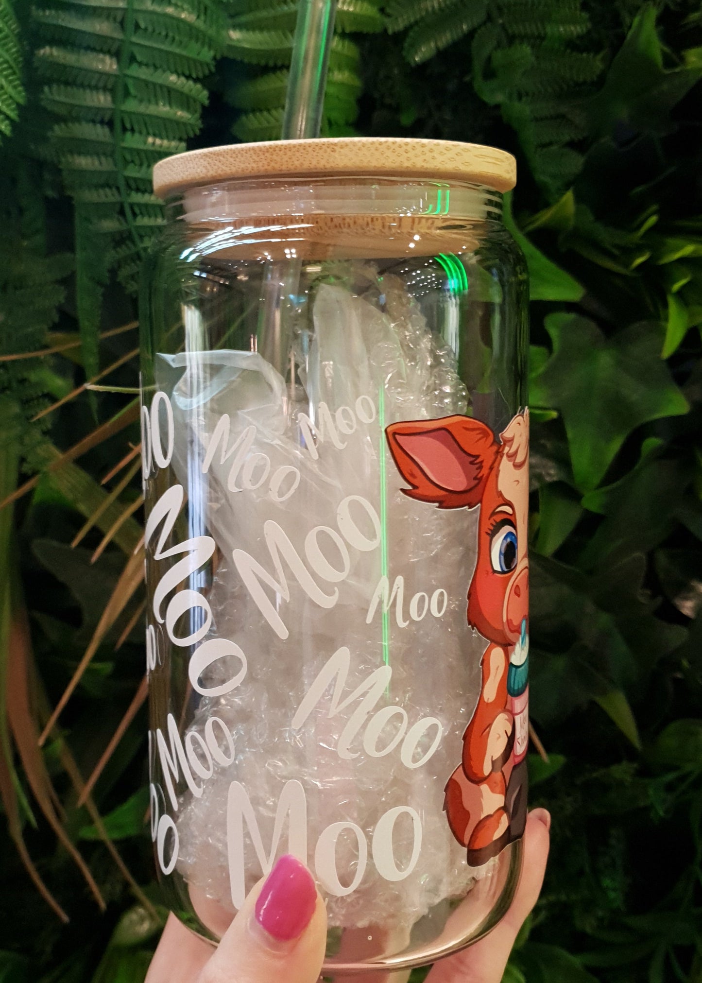 Clear tumbler with cow design and 'Moo' text held by a hand on green leaves