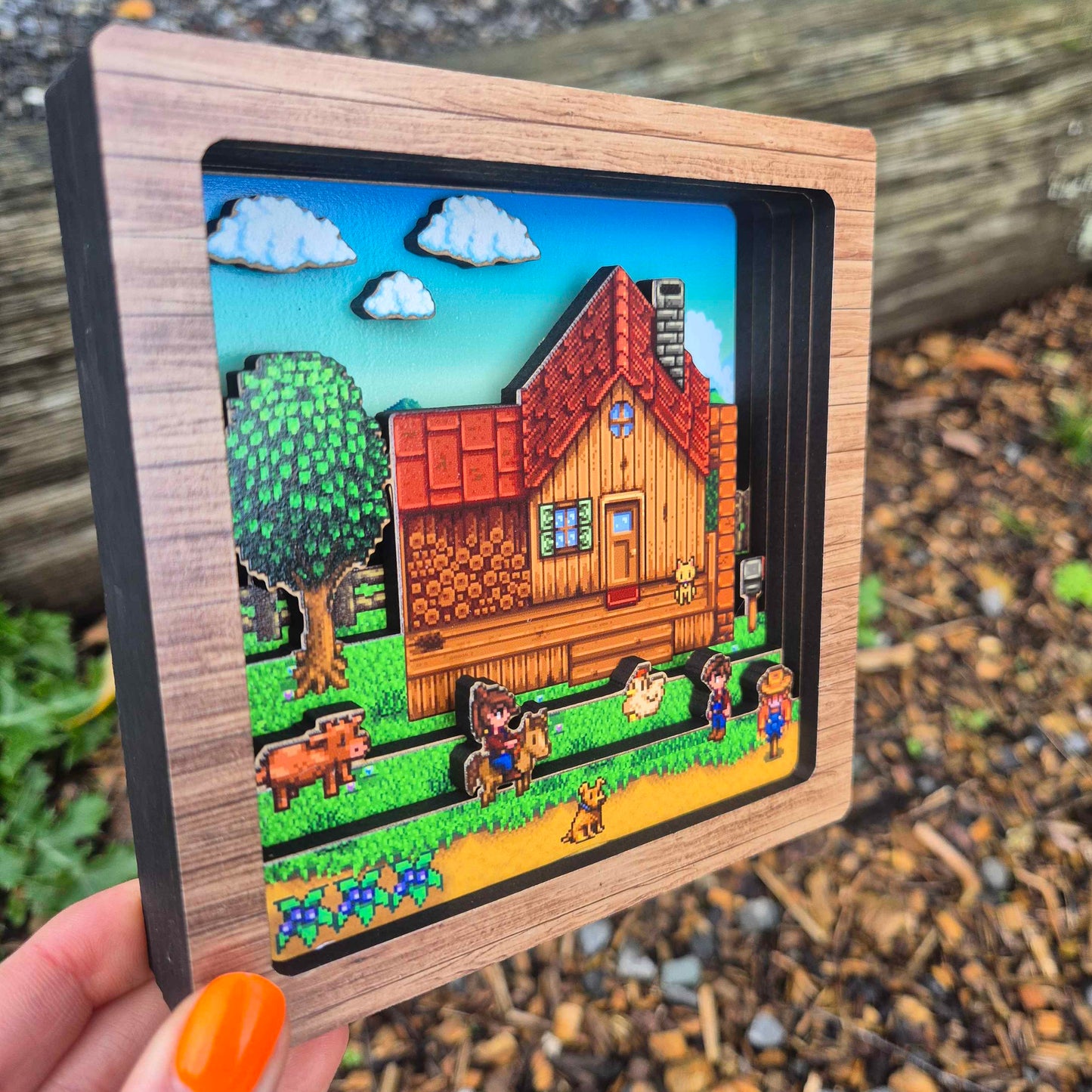 Hand holding a wooden-framed miniature landscape with a cabin and animals.