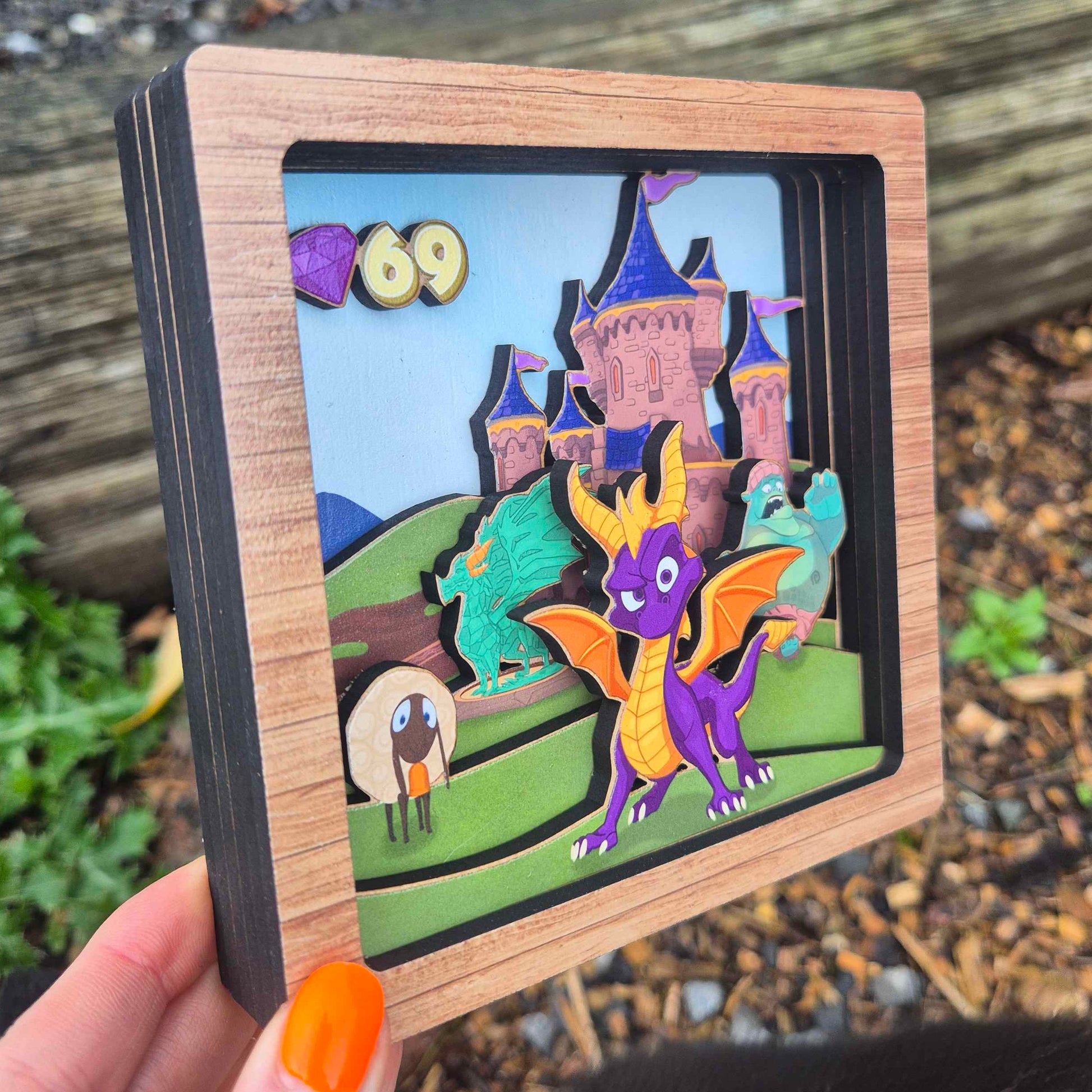Wooden box with a colorful illustration of a dragon and castle held by a hand outdoors.